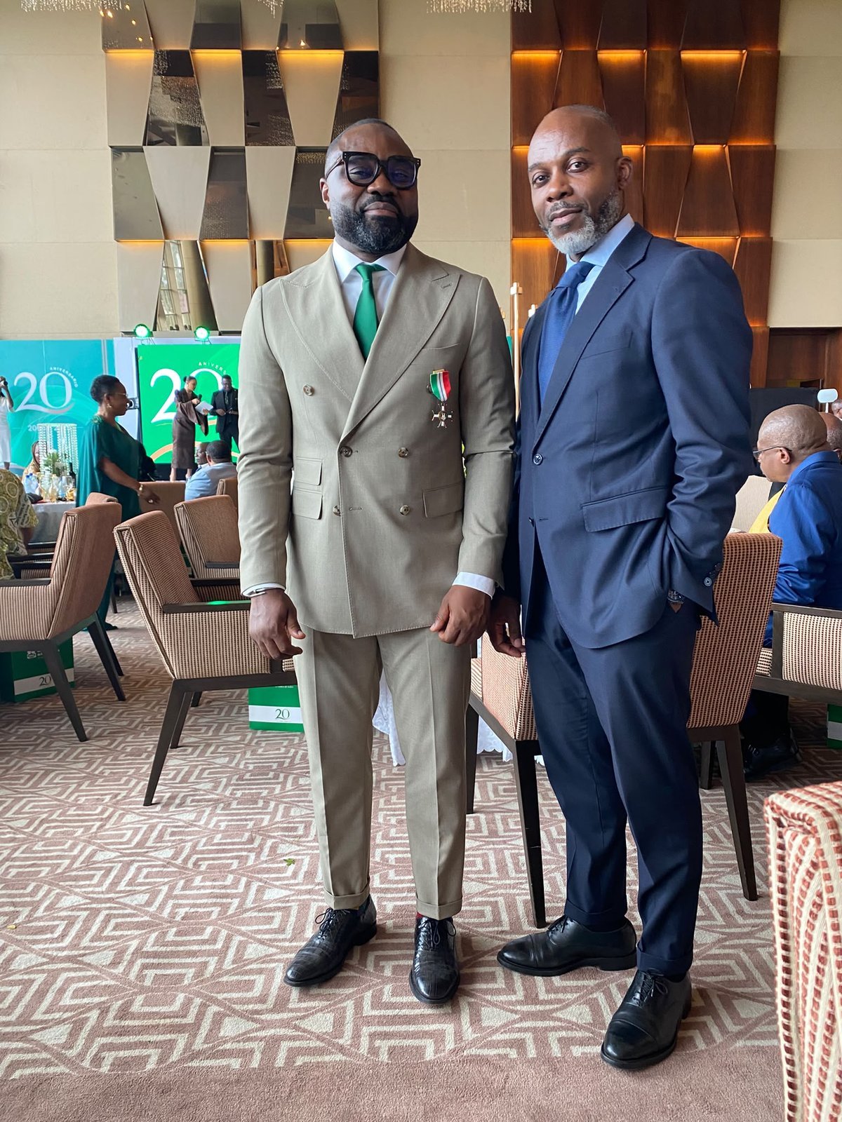 Two attendees pose in the hall, with the stage for BANGE's 20th anniversary in the background.